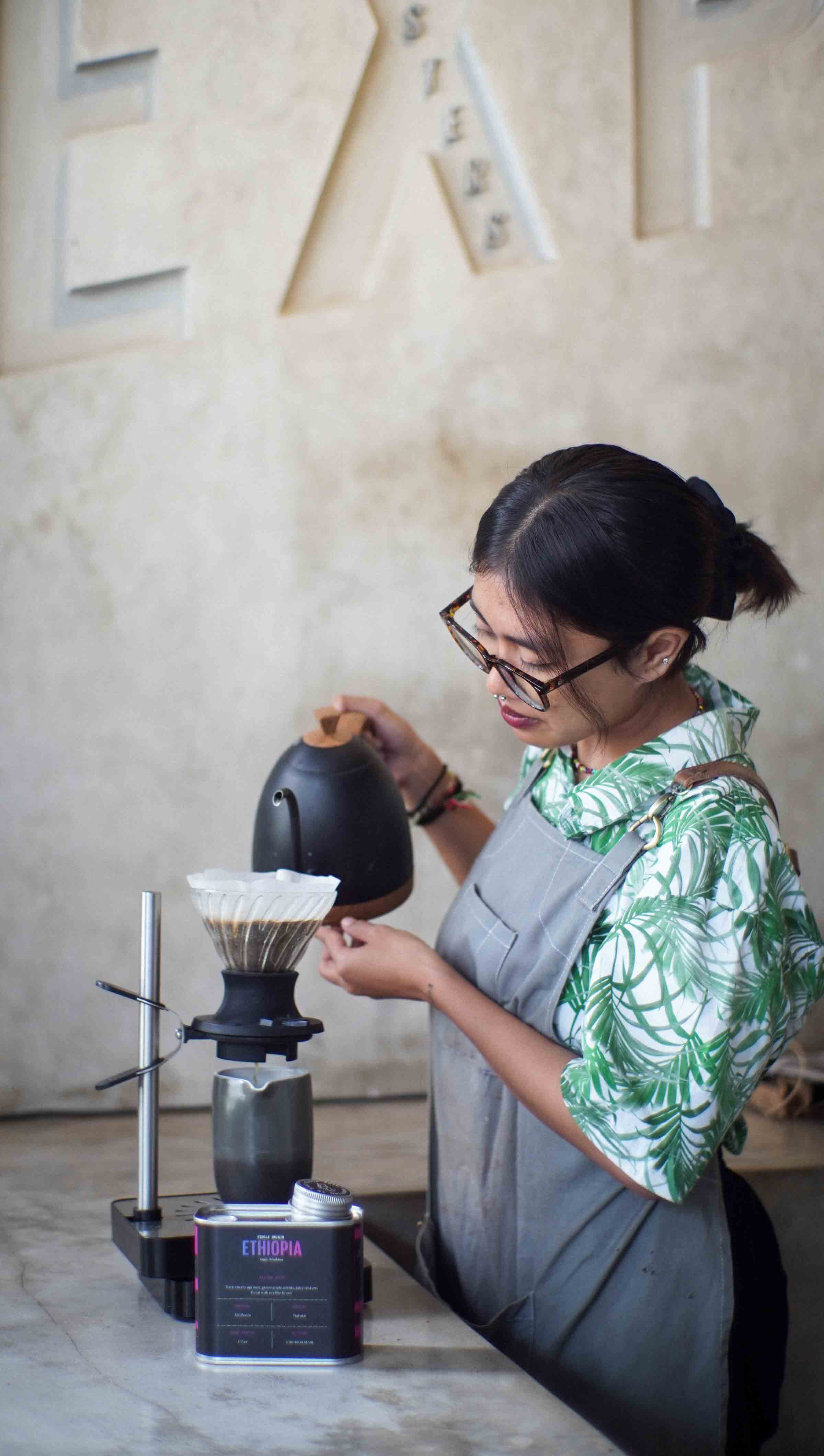 A barista in a tropical shirt carefully pouring hot water over a coffee filter in a pour-over coffee maker at Expat coffee roastery.