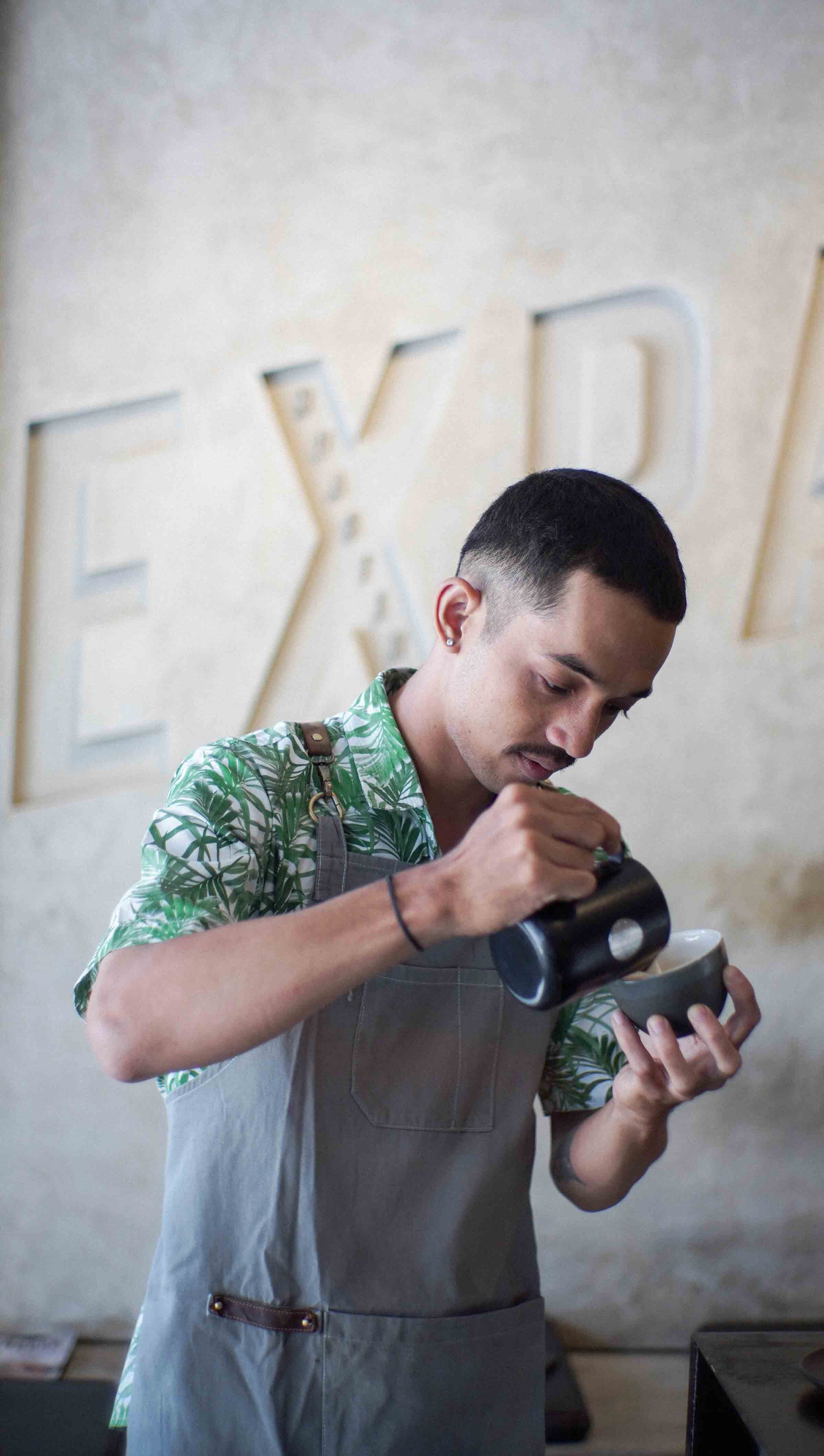 A barista in a tropical shirt pours steamed milk into a cup to create latte art, standing in front of the Expat coffee roastery sign.